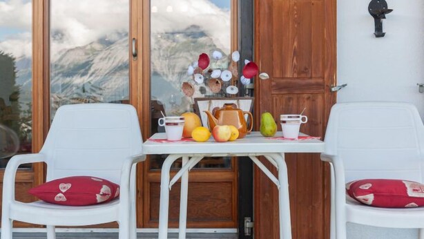 Een gedekte tafel met fruit en een theepot, twee witte stoelen met rode kussens, en een houten deur met uitzicht op bergen.