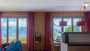 Een gezellige kamer met een blauwe bank, houten wanden, een klein houten kastje, een bureau met stoel en grote ramen met uitzicht op de bergen.
