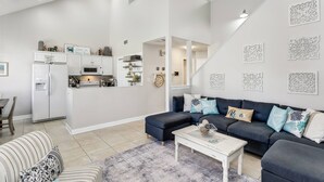A modern living room with a dark sofa, a white coffee table, and a striped armchair. A kitchen is visible in the background with white cabinets and a refrigerator.
