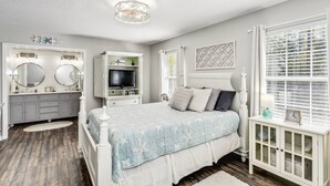 A bedroom with a bed, a vanity with mirrors, a television, and a window with blinds.