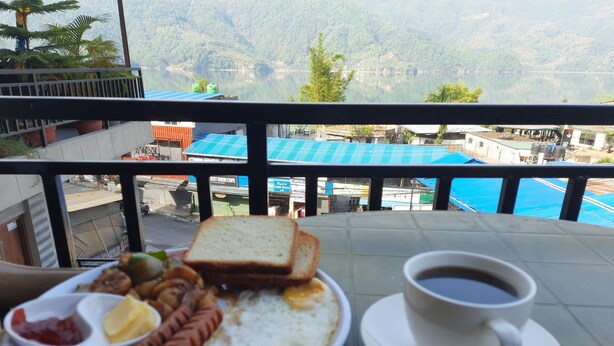 Un plato de desayuno con huevos, salchichas, tostadas y una taza de café en un balcón con vistas a un lago y edificios.