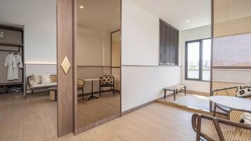 Een moderne hotelkamer met een houten vloer, een bank, een eettafel en een stoel. Er is een raam met zonwering en uitzicht op het omliggende landschap.