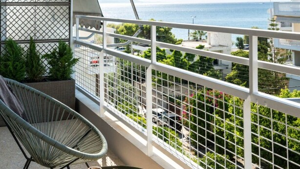 A balcony with a view of the sea, a chair, and a table with a glass of orange juice and cookies. - 2-BD Luxury Apt  Sea View Balcony near the beach (Voula)