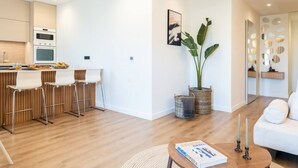 A modern living room with a wooden floor, a white sofa, a coffee table with a book, and a kitchen area with a white countertop and bar stools. - 2-BD Luxury Apt  Sea View Balcony near the beach (Voula)