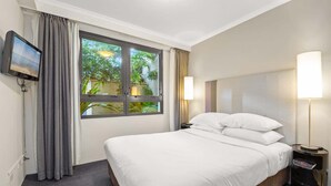 A hotel room with a bed, a TV mounted on the wall, a window with curtains, and a bedside lamp. - Contemporary 1-Bed First Floor Unit with Pool (Parramatta)