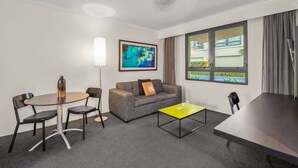 A hotel room with a grey sofa, a yellow coffee table, a round dining table with chairs, a floor lamp, and a large window with curtains. - Contemporary 1-Bed First Floor Unit with Pool (Parramatta)