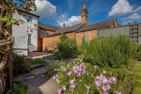 A traditional stone building with a brick chimney, a white exterior wall, and a garden with purple flowers.