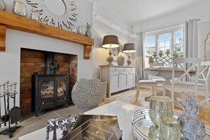 A cozy living room with a fireplace, white cabinets, a wooden shelf with decorative items, and a dining area with white chairs and a table.
