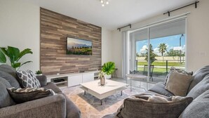 A modern living room with a flat-screen TV mounted on a wooden wall, a white coffee table, grey sofas, and a large sliding glass door leading to an outdoor patio. - Superb, Spacious Condo, Prime Resort! (Kissimmee)