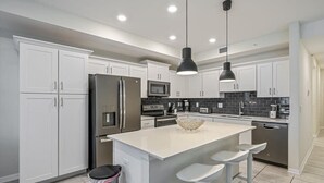 A modern kitchen with white cabinets, stainless steel appliances, a central island with bar stools, and pendant lighting. - Superb, Spacious Condo, Prime Resort! (Kissimmee)