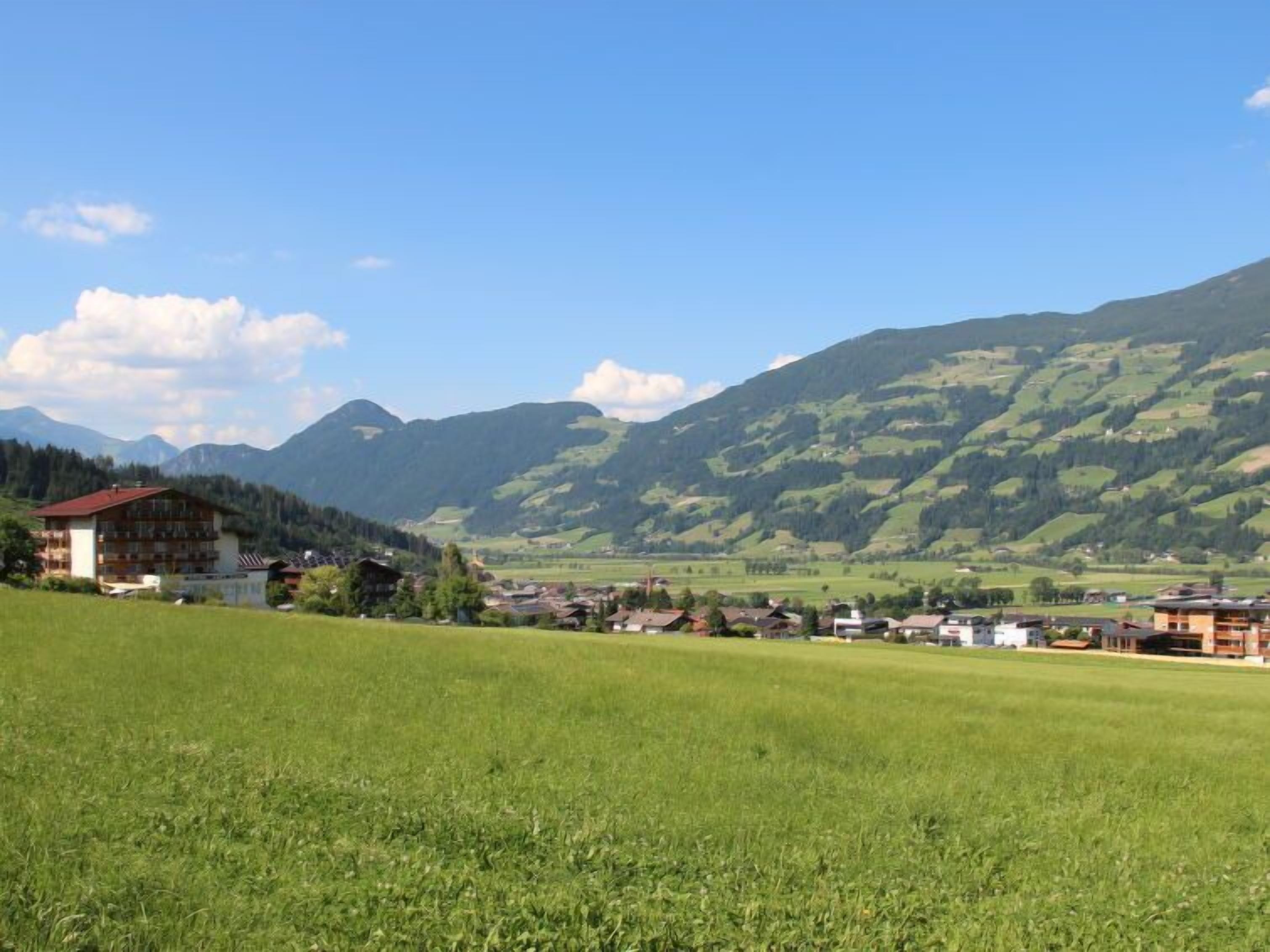 A rural landscape with a building that appears to be a hotel or guesthouse, surrounded by green fields and mountains in the background.