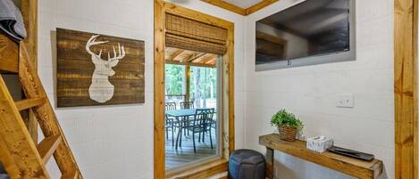 Una habitación con una escalera de madera, un televisor montado, una obra de arte con cabeza de venado enmarcadas y una puerta que da a una zona de estar exterior.