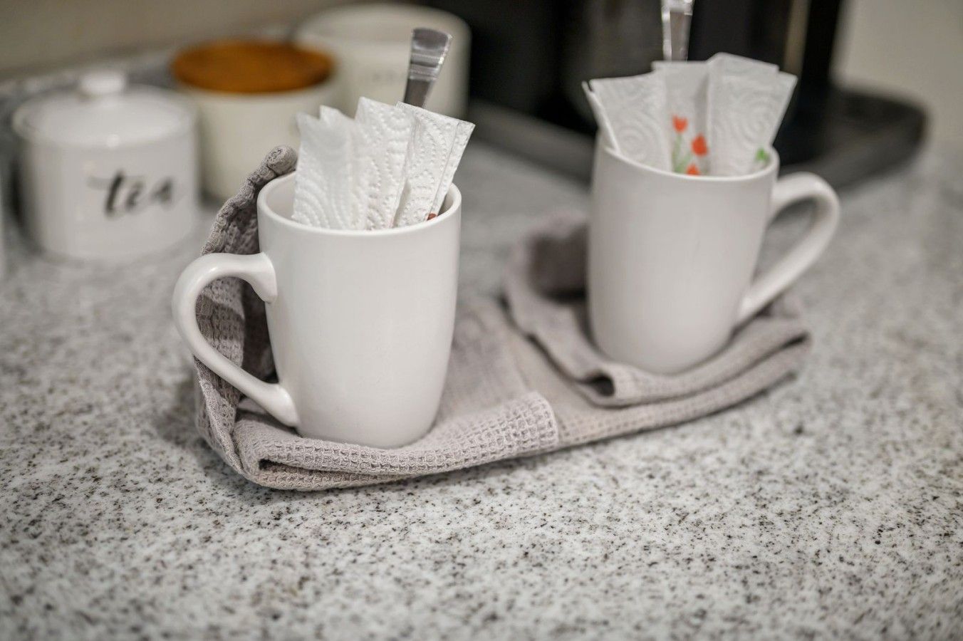 Two white ceramic mugs on a speckled countertop, each with a napkin holder containing a napkin.