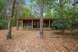 A cabin with a porch, surrounded by trees and a forest floor covered with fallen leaves. - EZ Ridge Retreat | Rustic Broken Bow Lake Cabin with Hot Tub, Fire Pit, Grill & Pets (Hochatown)