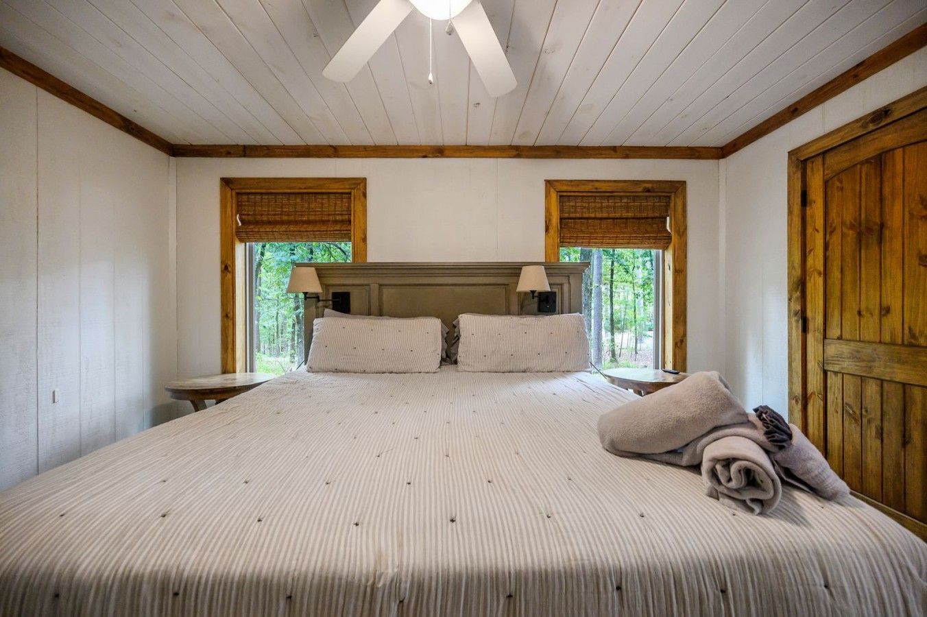 Un dormitorio con una cama grande, dos mesitas de noche, un ventilador de techo y ventanas con contraventanas de madera.