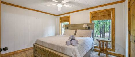 Un dormitorio con cama, ventilador de techo, ventana con vista a árboles y una pequeña mesa.