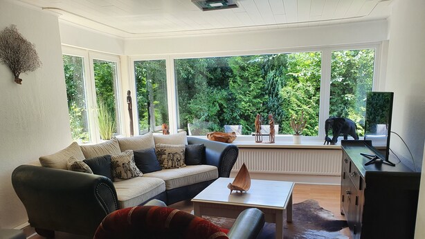 A modern living room with a sofa, a coffee table, and a red armchair. There is a large window with a view of greenery outside.