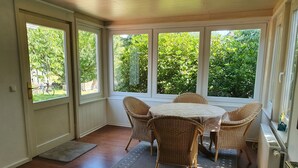 A sunlit room with a round table and wicker chairs, large windows, and a radiator.