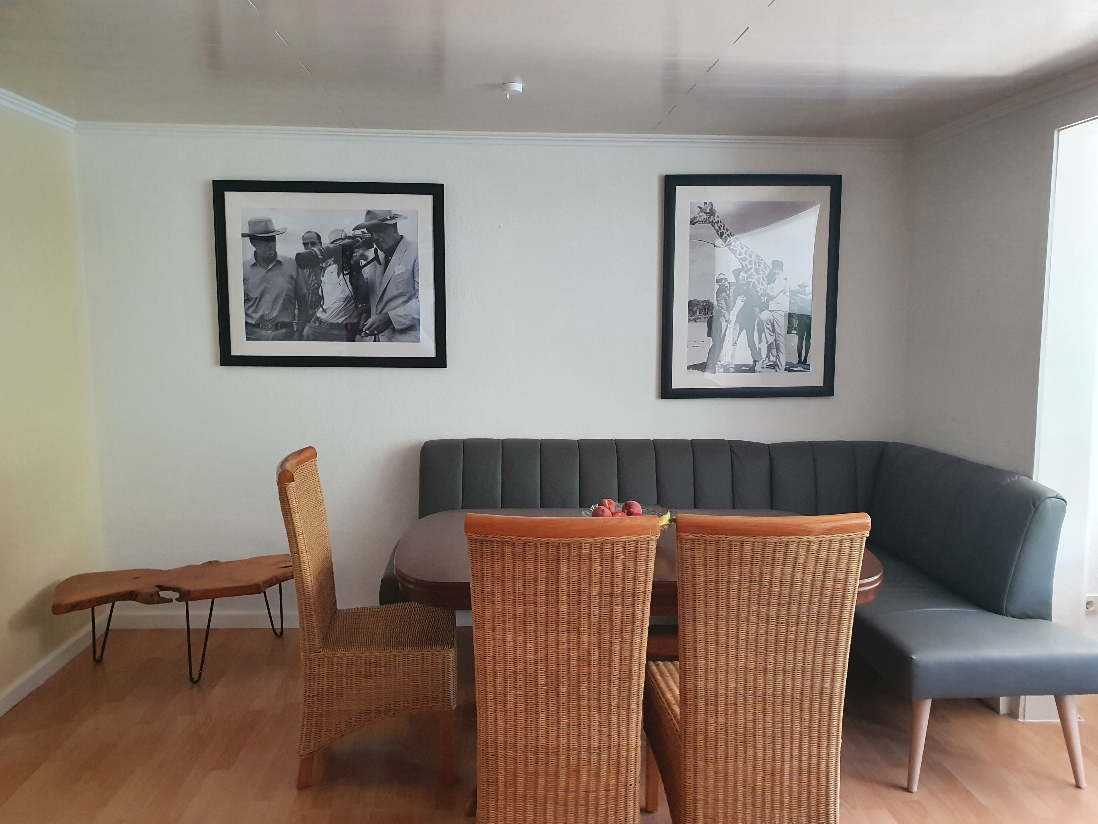 Een woonkamer met een grijze bank, rieten stoelen, een klein tafeltje en twee ingelijste foto’s aan de muur.