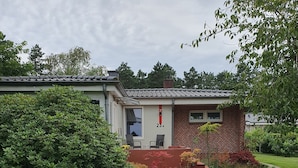 A single-story house with a brick exterior, a visible number '23' on the wall, a small porch with two chairs, and a well-maintained lawn with a concrete pathway leading to the house.