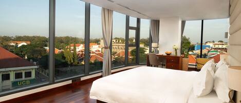 A hotel room with a large bed, a desk with a chair, and a view of the cityscape through large windows.