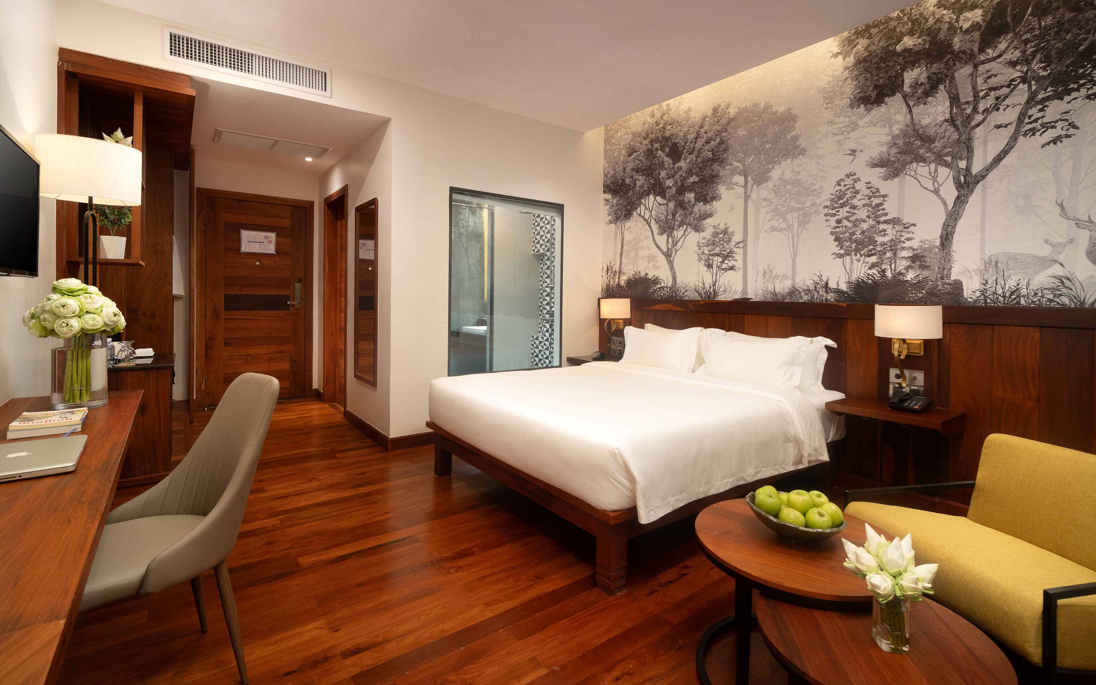 A hotel room with a bed, a desk, a chair, a small table with apples and flowers, and a wall with a nature mural.