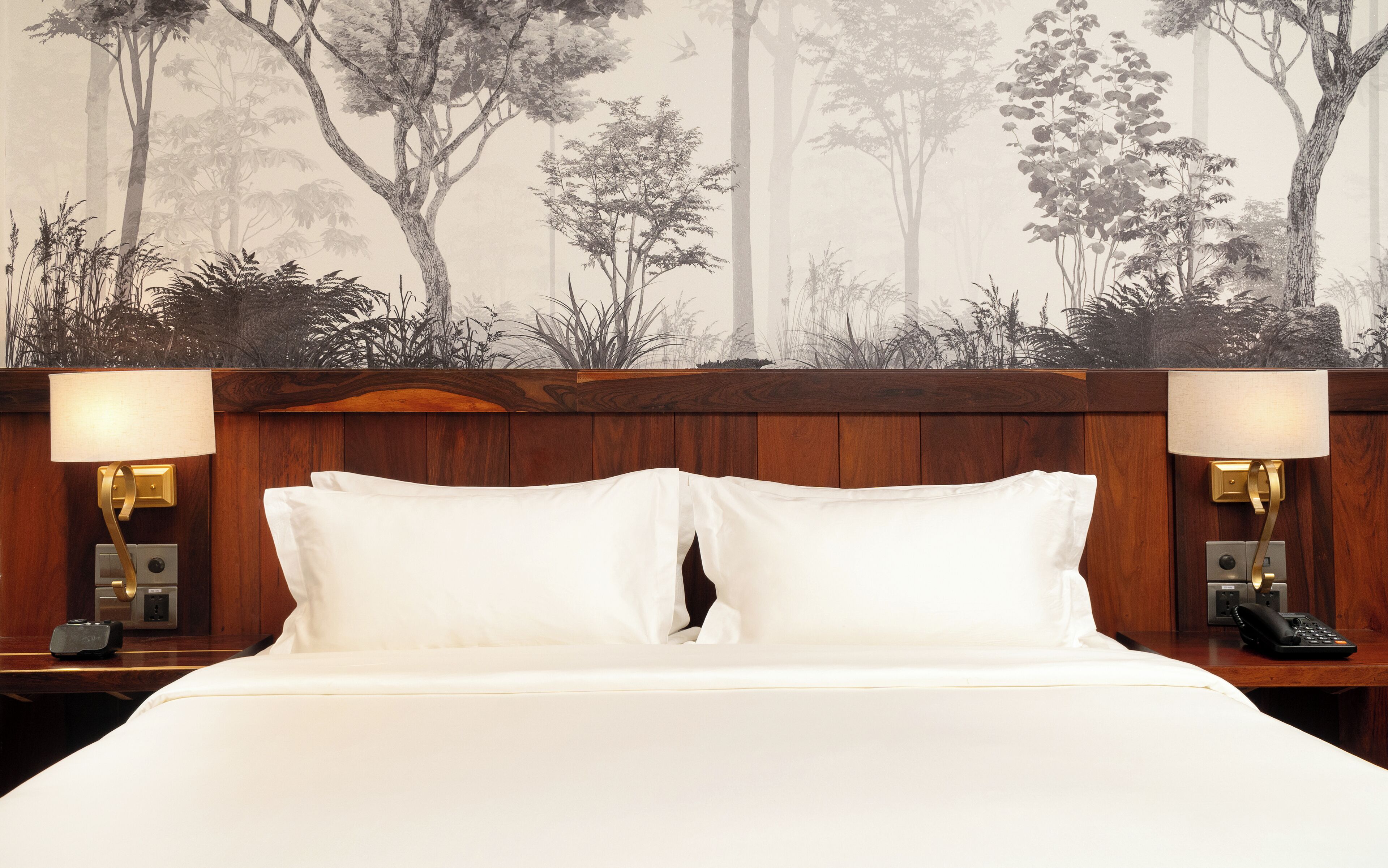 Habitación de hotel con una cama grande, dos lámparas de noche, un cabecero de madera y un mural de árboles en la pared.
