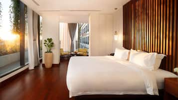 Habitación de hotel moderna con una cama grande, una planta en maceta, una mesita con un frutero y vistas a la ciudad a través de amplias ventanas.
