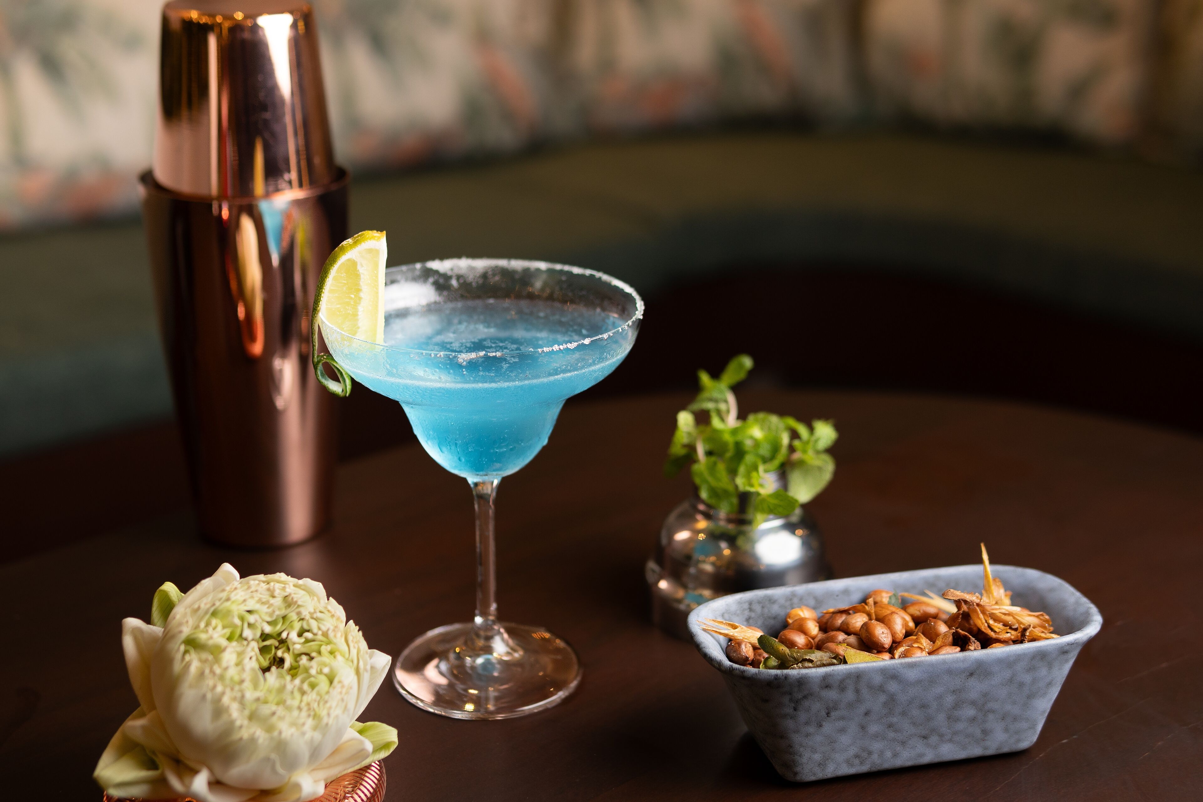 A cocktail glass with a blue drink, a copper shaker, a flower, and a bowl of nuts on a table.