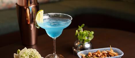 A cocktail glass with a blue drink, a copper shaker, a flower, and a bowl of nuts on a table.