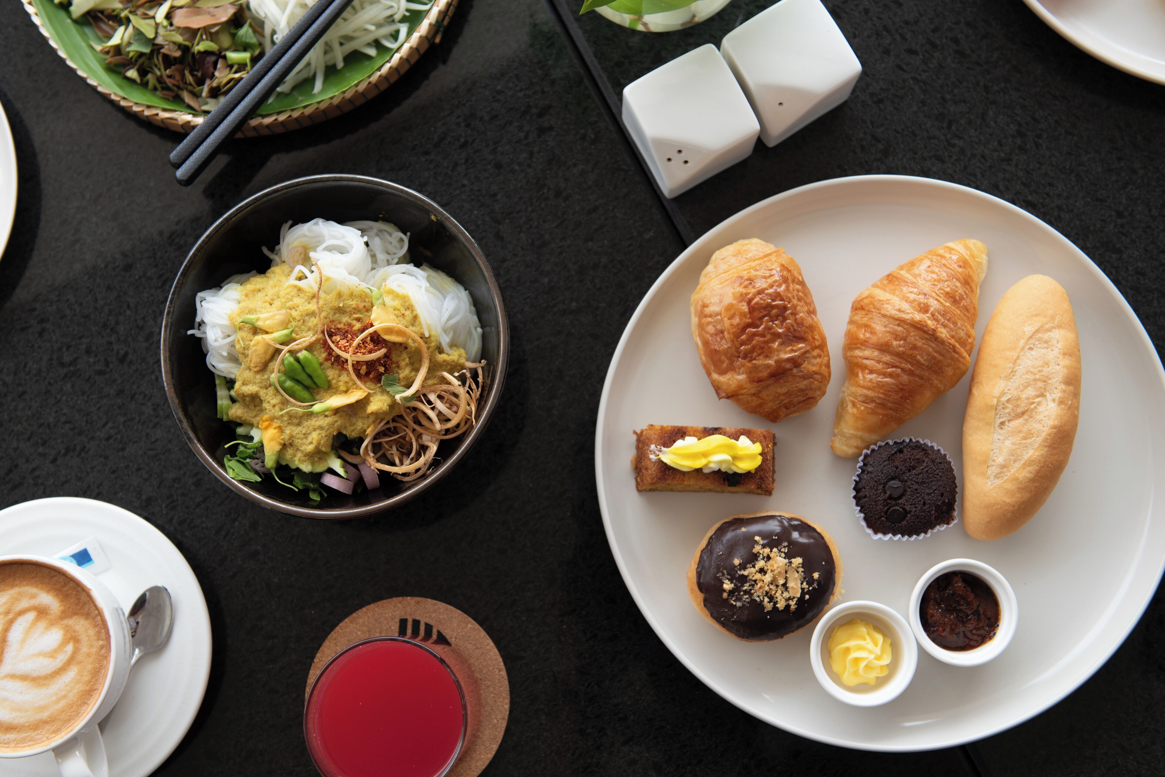 A table with a variety of food items including a bowl of noodles, croissants, cupcakes, and a cup of coffee.