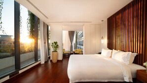 Une chambre d’hôtel moderne dotée d’un grand lit, d’un petit coin salon avec une table et des chaises, et d’une grande fenêtre donnant sur la ville.