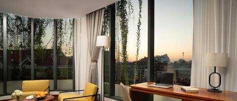 Habitación de hotel moderna con un escritorio de madera, dos sillas, una computadora portátil y vistas a la ciudad al atardecer.