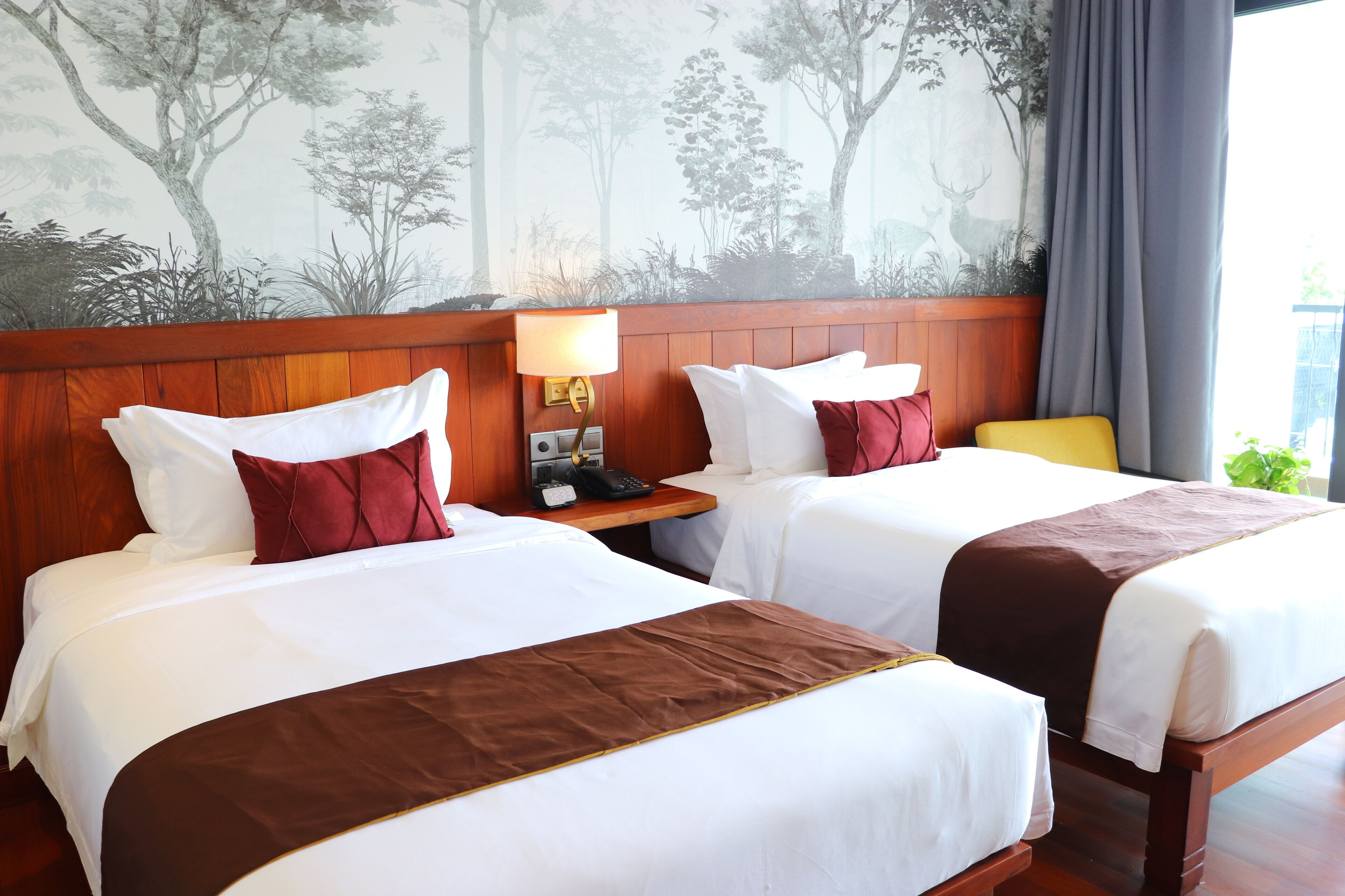 Habitación de hotel con dos camas, cabecera de madera, mesita de noche con lámpara y un mural de árboles en la pared.