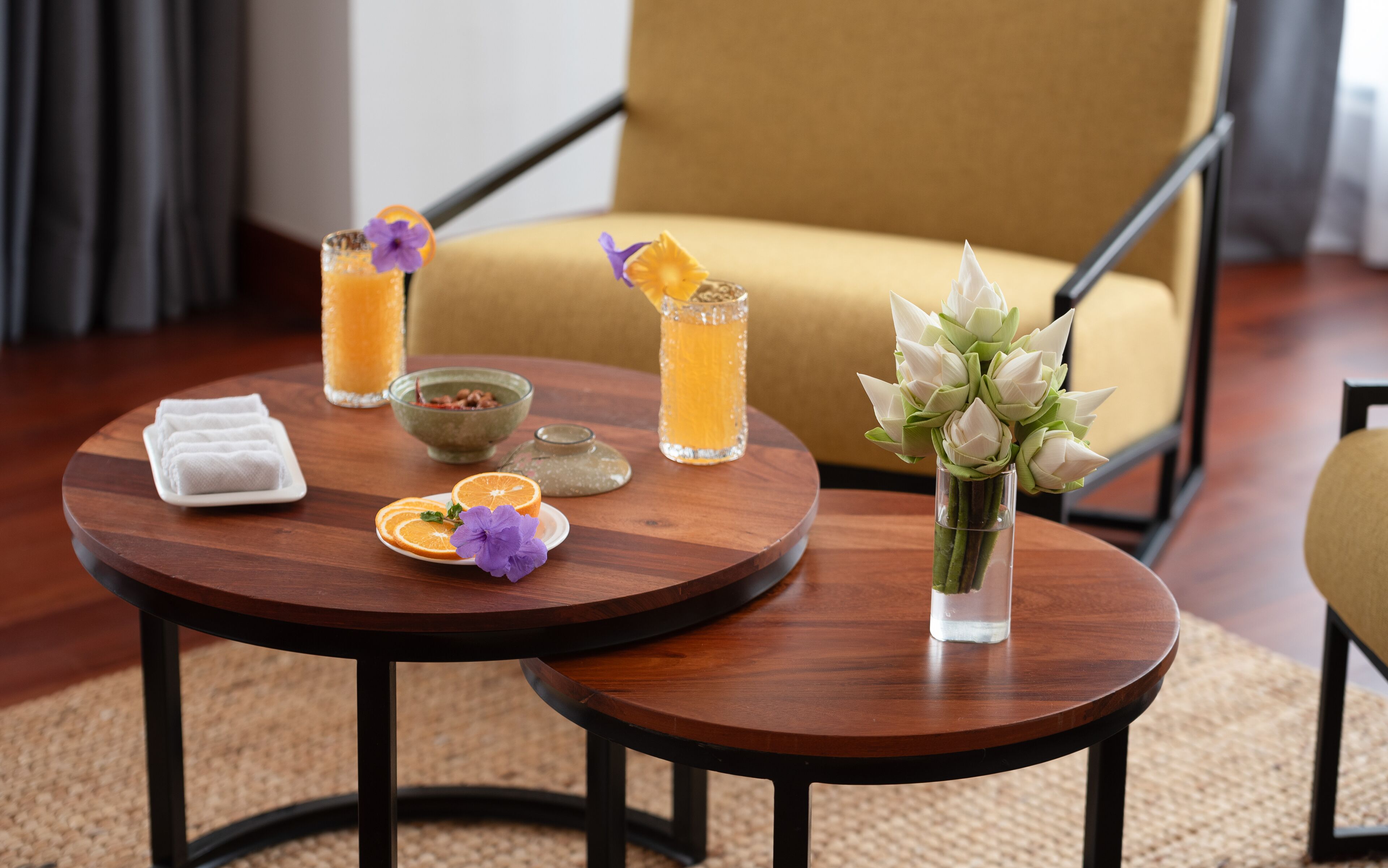 A round wooden table with a vase of white flowers, a glass of orange juice, a bowl of food, and folded towels.