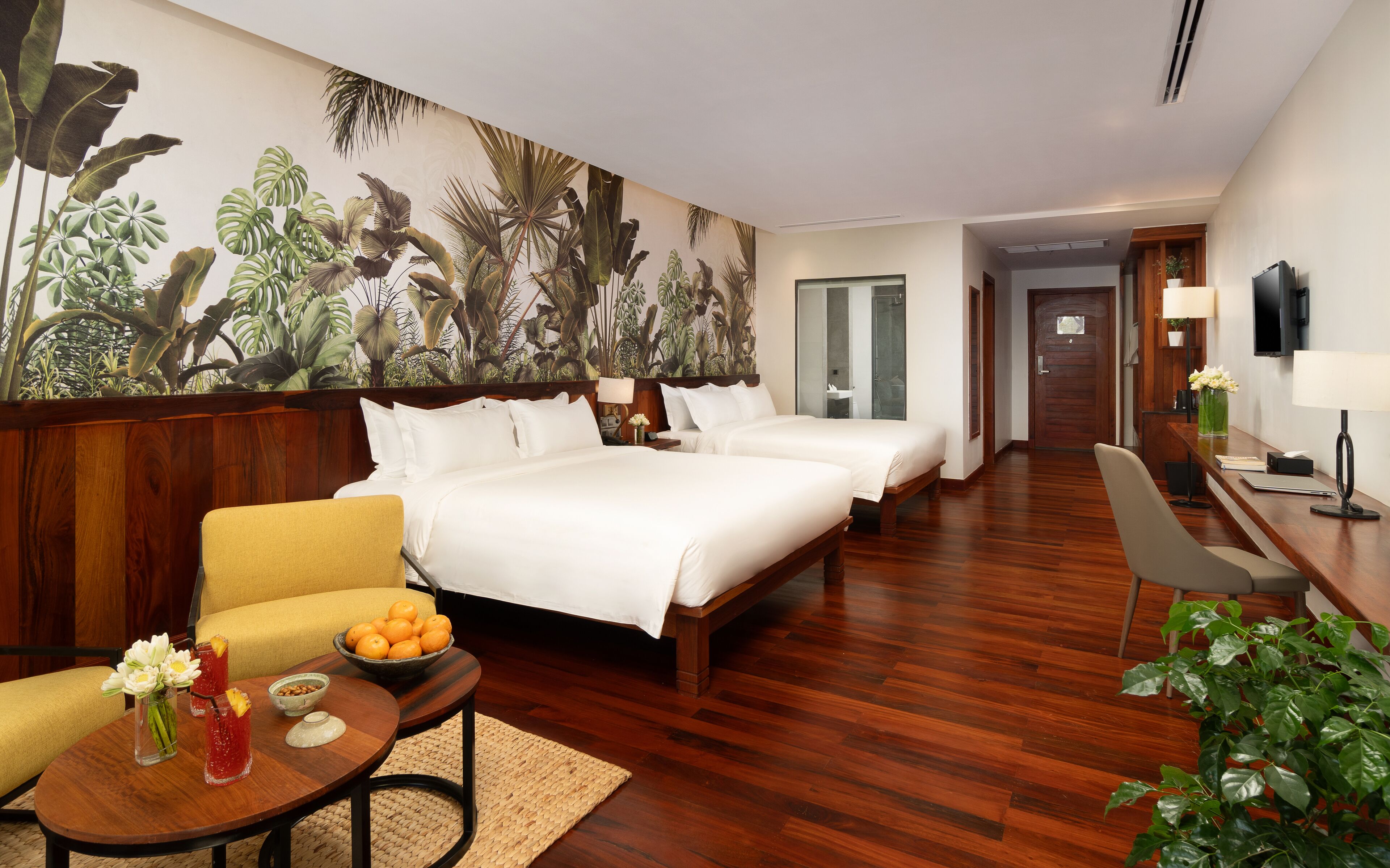 Habitación de hotel con un amplio mural tropical, dos camas, un escritorio de madera, una silla, una pequeña mesa con fruta y bebidas, y una planta en maceta.