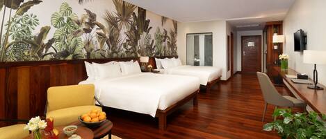 Habitación de hotel con un amplio mural tropical, dos camas, un escritorio de madera, una silla, una pequeña mesa con fruta y bebidas, y una planta en maceta.