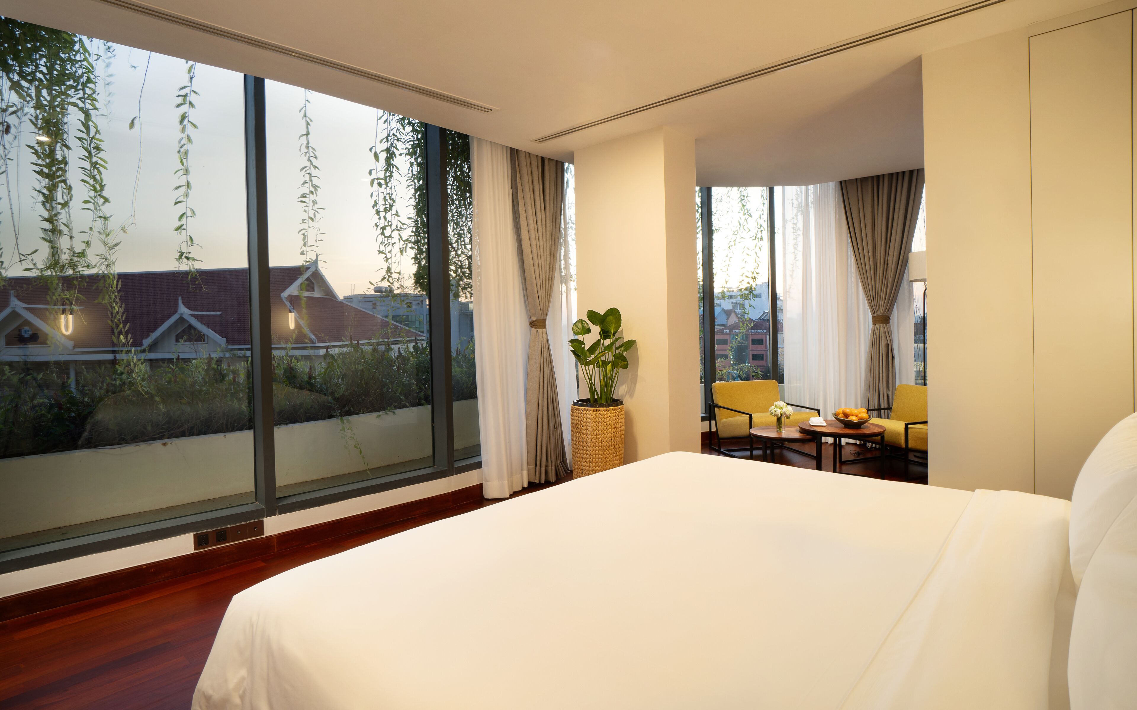 A hotel room with a large bed, a small table with a vase and fruit bowl, two chairs, and a view of buildings through large windows.