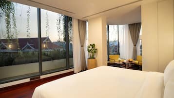 A hotel room with a large bed, a small table with a vase and fruit bowl, two chairs, and a view of buildings through large windows.