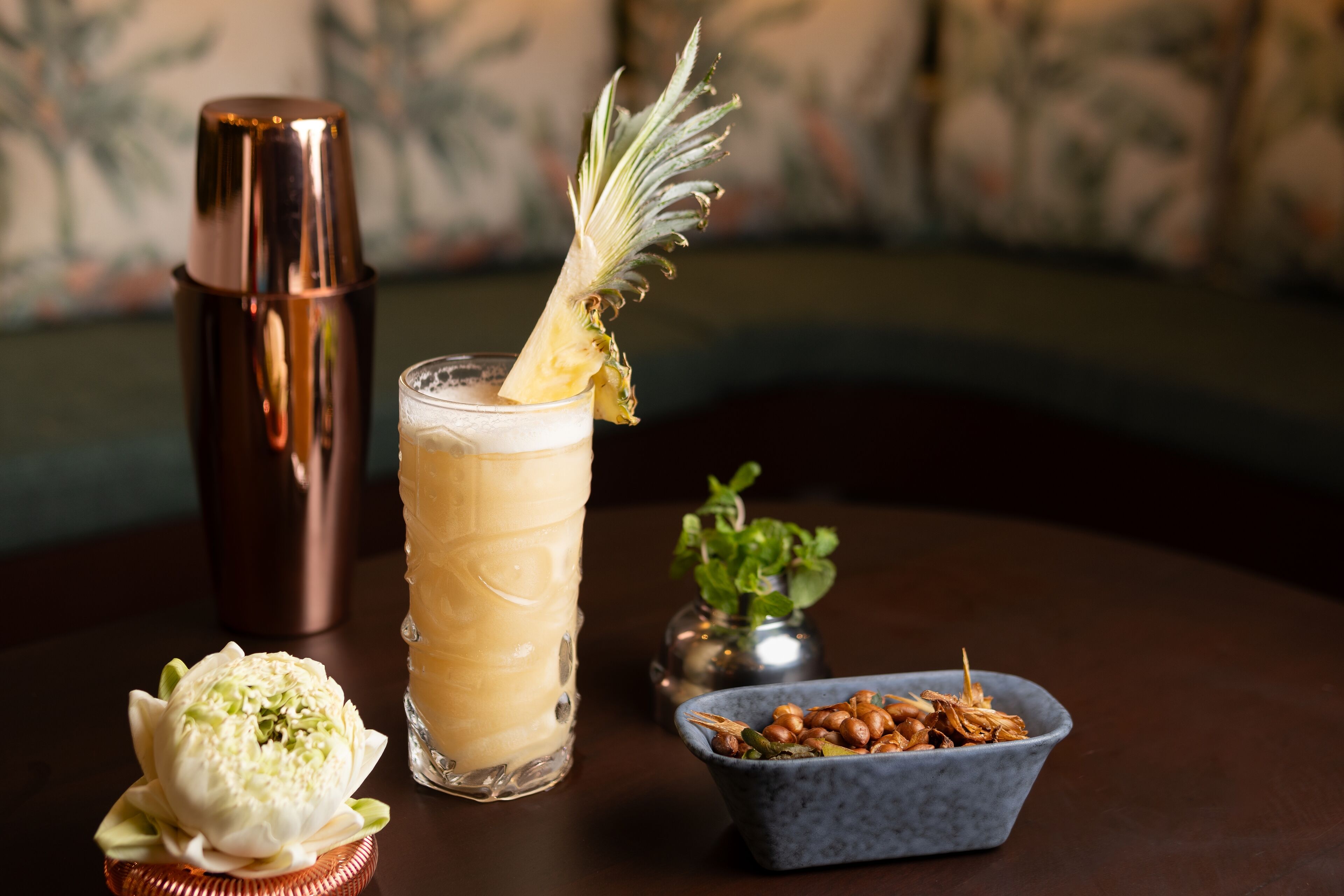 A cocktail glass with a pineapple garnish, a copper cocktail shaker, a bowl of nuts, and a flower on a table.