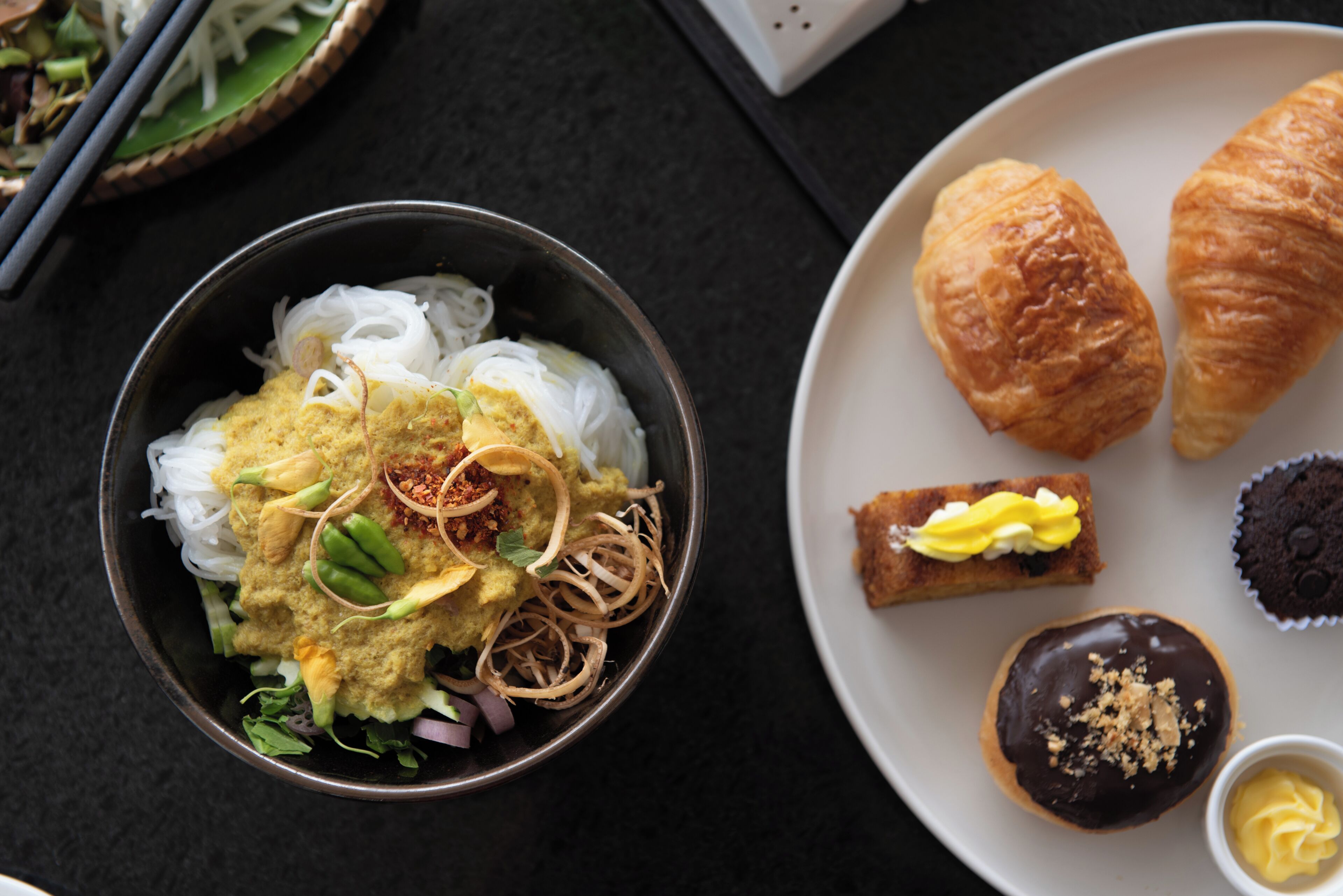 A bowl of noodles with toppings, a plate with croissants and pastries, and a pair of chopsticks on a dark surface.