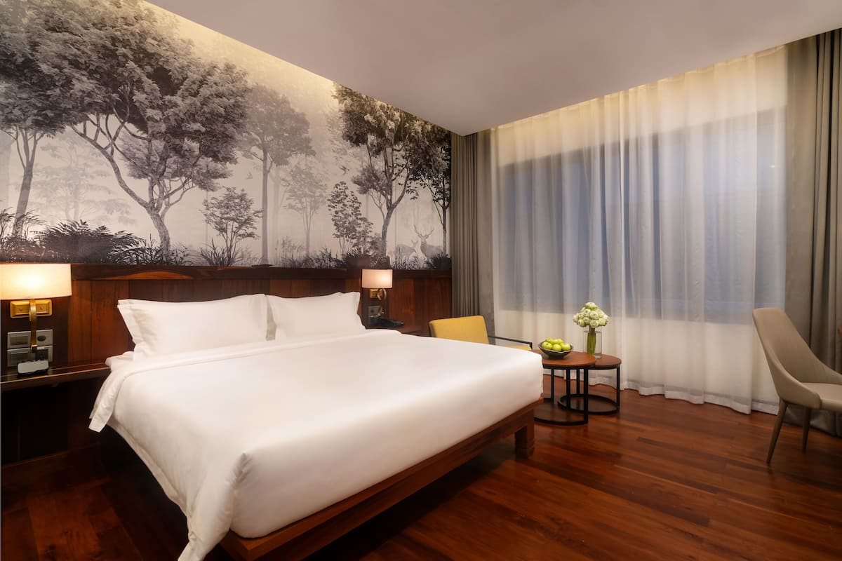 Hotelkamer met een groot bed, een houten hoofdeinde, een nachtkastje met een lamp, een tafeltje met een bloemenvaas, een stoel en een bureau met een lamp.