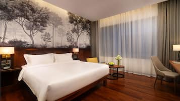 Une chambre d’hôtel avec un grand lit, une tête de lit en bois, une table de chevet avec une lampe, une petite table sur laquelle est posé un vase de fleurs, une chaise et un bureau avec une lampe.