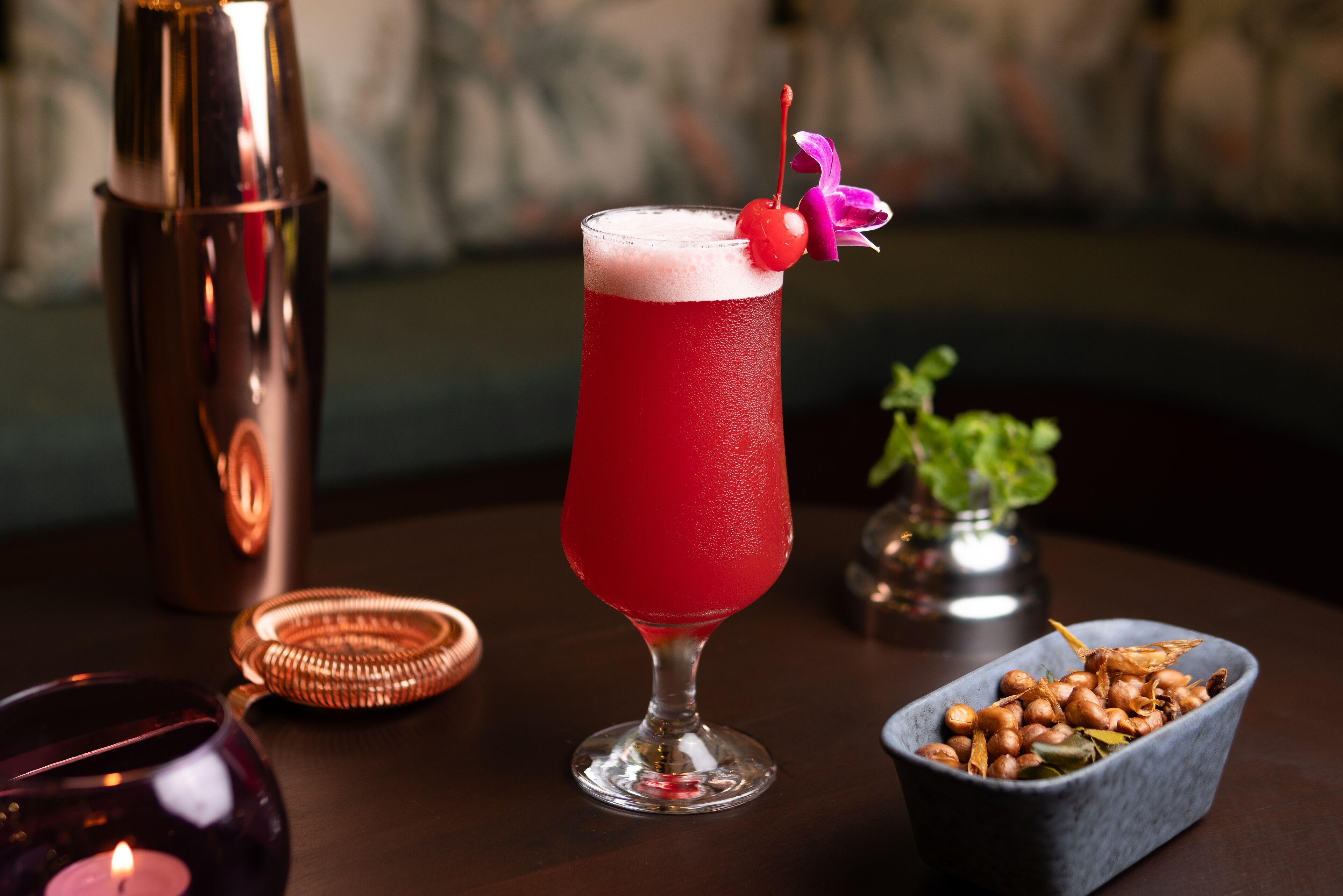 A cocktail in a tall glass with a cherry garnish, a candle, and a small plant on a table.