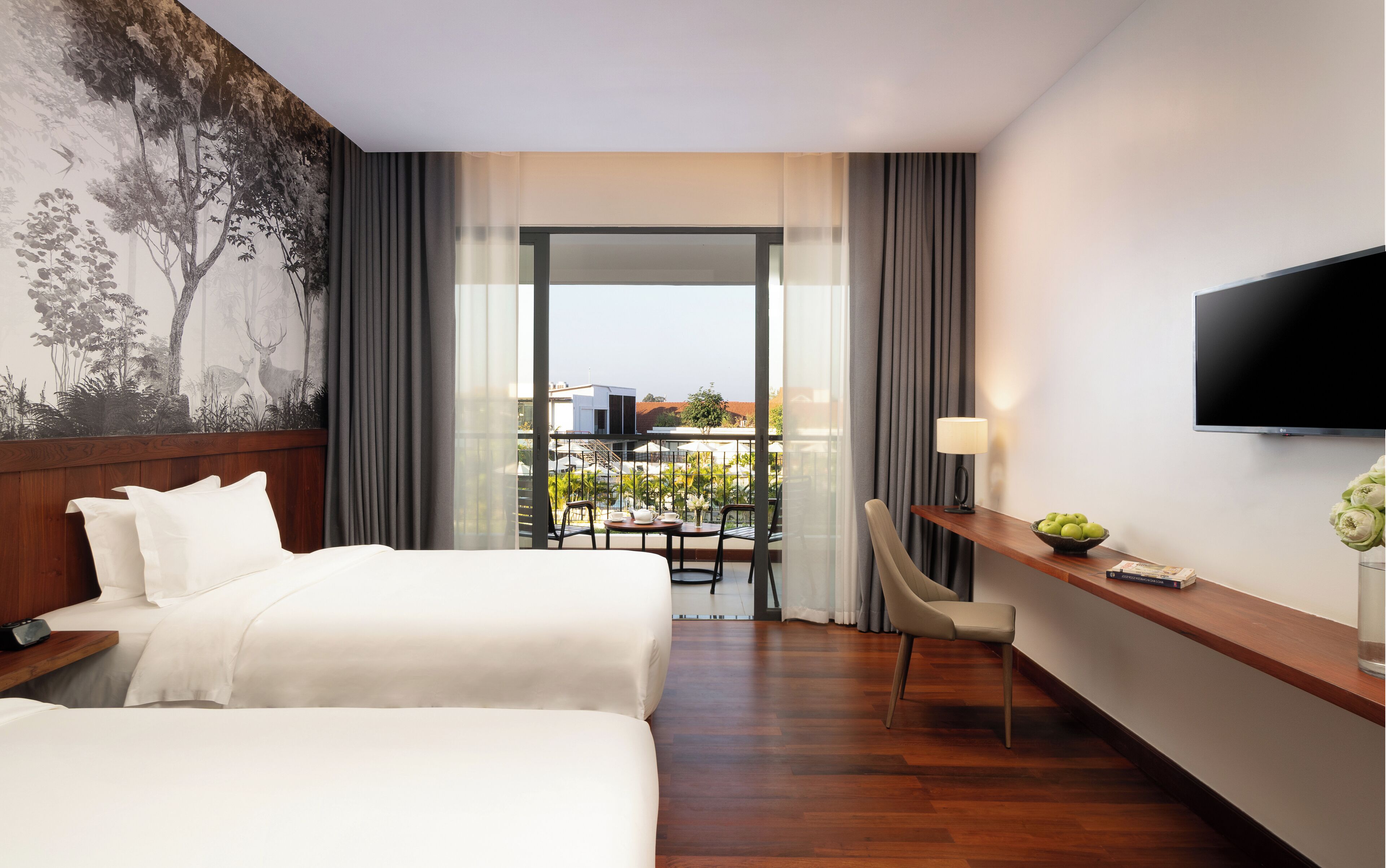 A hotel room with two beds, a desk with a lamp, a TV, and a balcony with a view of buildings.