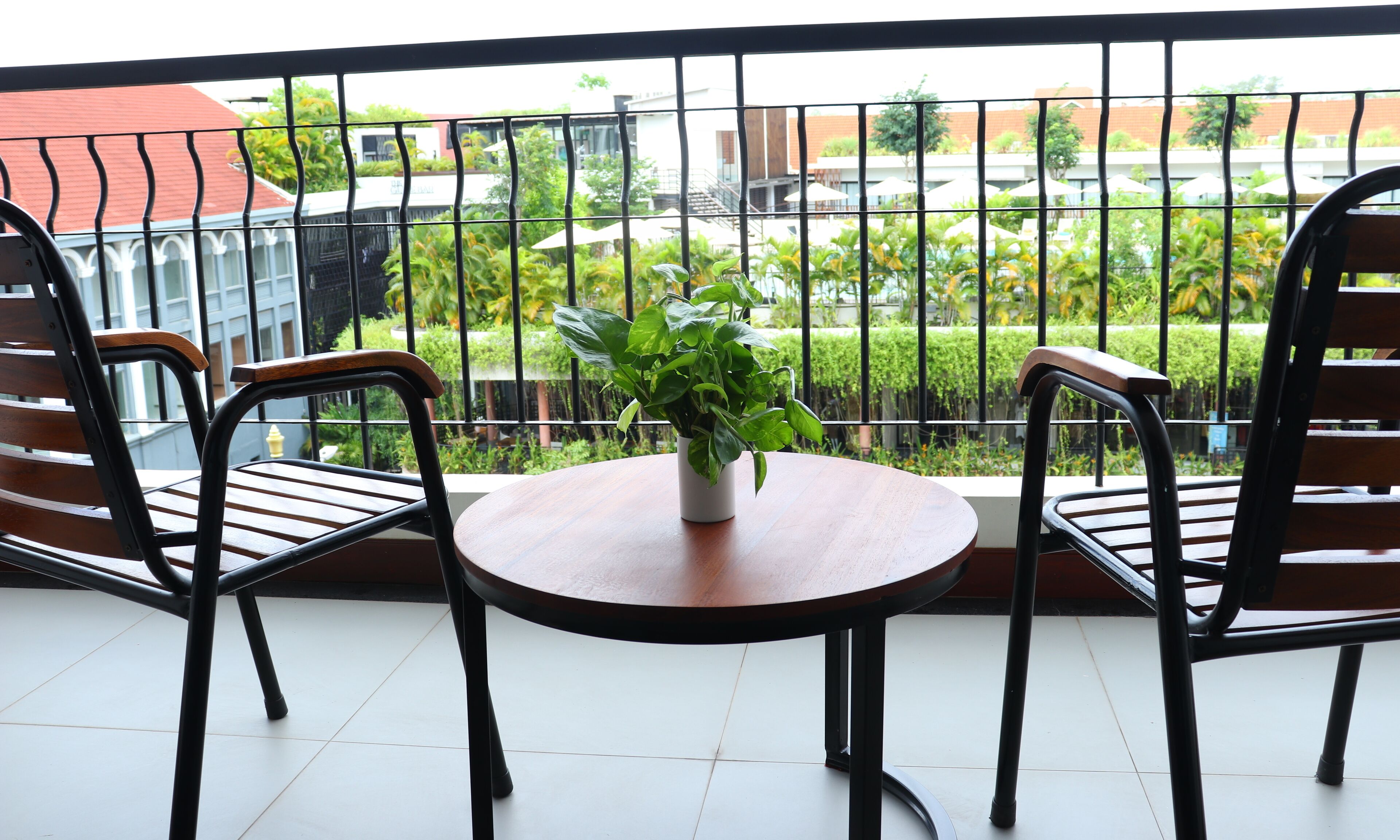 Un balcon avec deux chaises en bois et une petite table, une plante en pot, et une vue sur un jardin et des bâtiments.