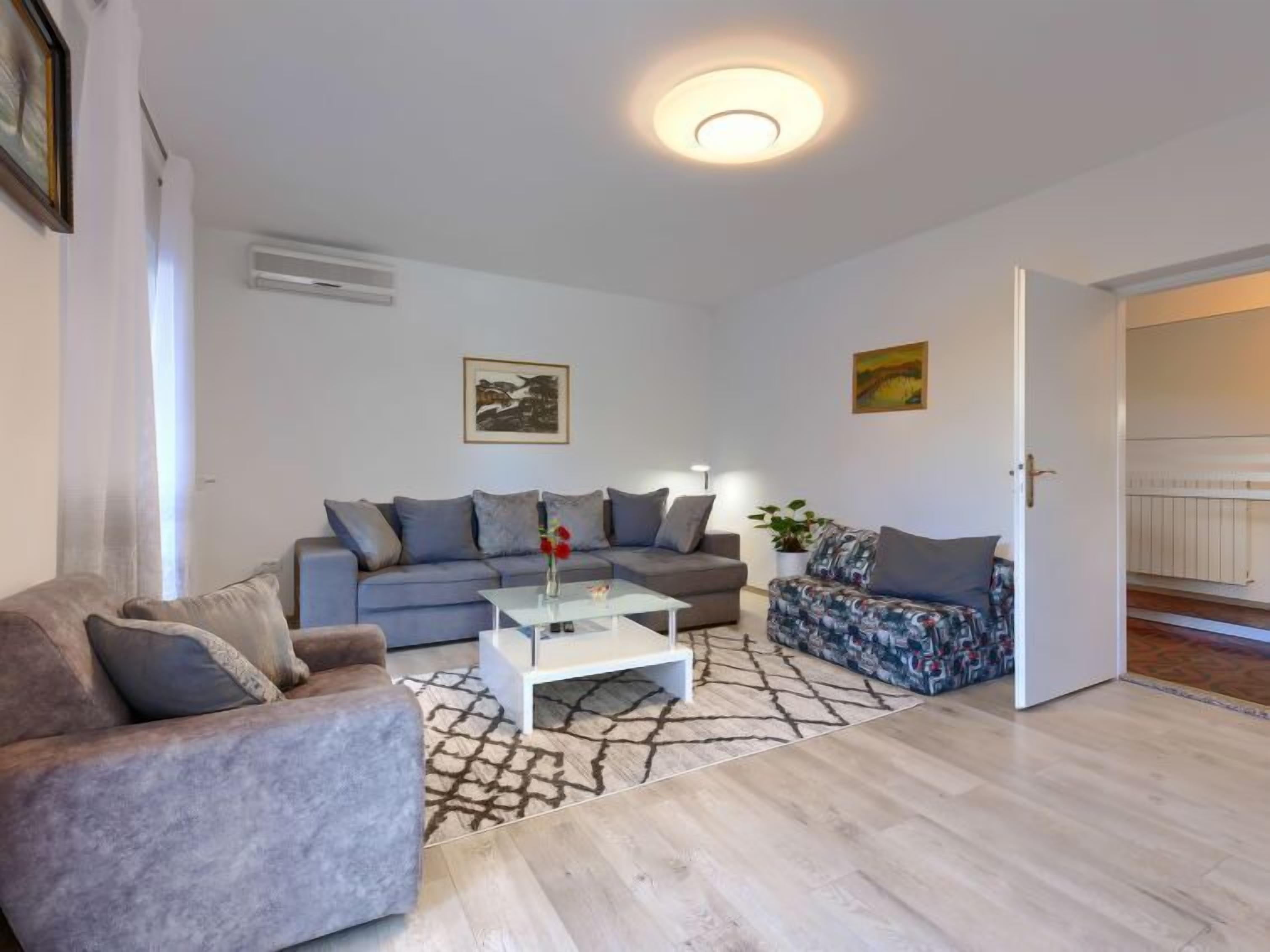 Een moderne woonkamer met een grijze bank, een gestreept bankstel en een salontafel. Er hangt een schilderij aan de muur en een deur naar een andere kamer.