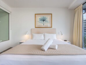 Una cama bien hecha con una toalla enrollada, dos lámparas de noche, una obra de arte enmarcada sobre la cama y una ventana con cortinas a la derecha.