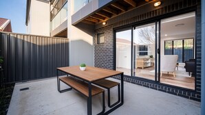 An outdoor patio with a wooden table and bench, a brick wall, and a sliding glass door leading to an interior space with furniture. - Elegance and Style (Wagga Wagga)