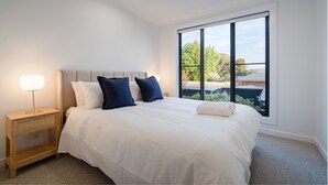 A neatly made bed with white and blue bedding, a bedside lamp, and a large window with a view of buildings and trees. - Elegance and Style (Wagga Wagga)